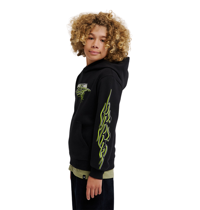 A young person with curly hair wears the DC Shoes Side Flame Hoodie Youth, a black fleece pullover featuring neon yellow-green DC graphics on the sleeve and chest, standing against a plain white background.