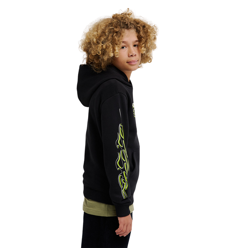 A youth model with curly blonde hair wears the DC Shoes Side Flame Hoodie, a black fleece featuring neon green flame designs on the sleeve, standing sideways and looking at the camera against a plain white background.
