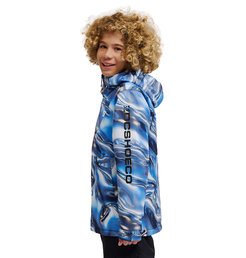 A smiling child with curly blond hair wears the DC Shoes Basis Youth Jacket—an insulated youth jacket featuring a blue, black, and white pattern and DCSHOECO text on the sleeve—standing sideways and looking at the camera.