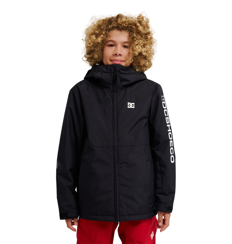 A young person with curly blond hair stands against a white background, wearing the DC Shoes Basis Youth Jacket—a black, durable, and stylish hooded jacket with DCSHOECO printed on the left sleeve—paired with red pants.