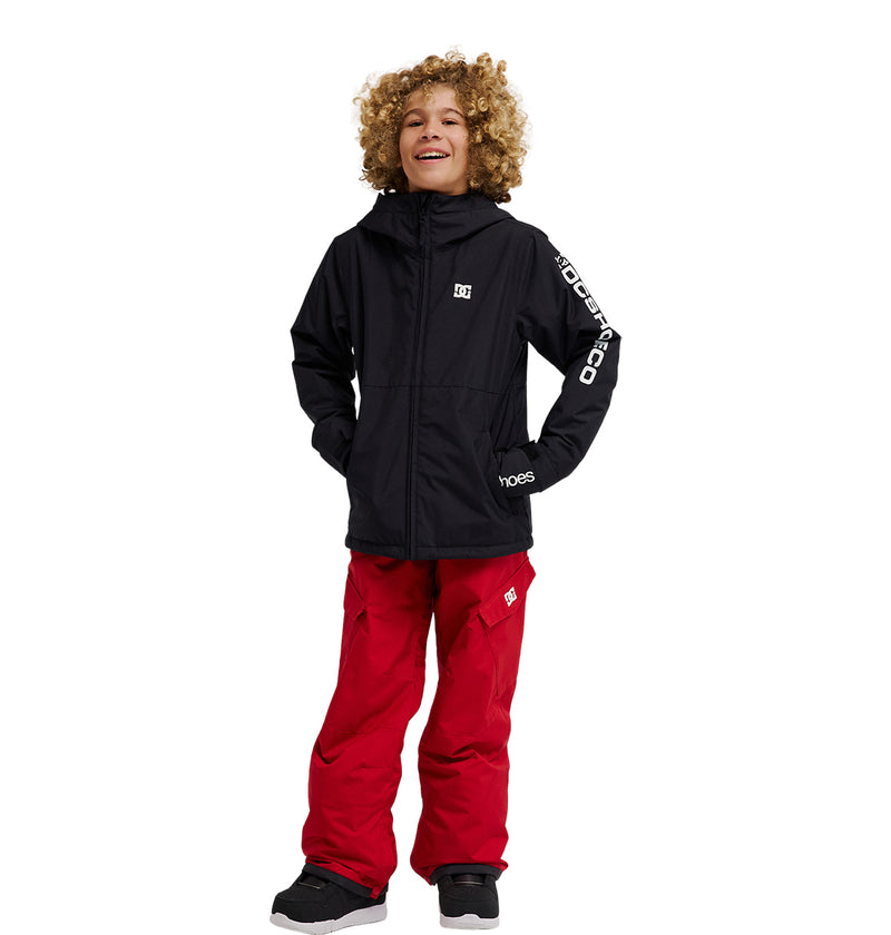 A smiling child with curly hair wears the DC Shoes Basis Youth Jacket, featuring white lettering on the sleeve, paired with bright red snow pants and standing with hands in jacket pockets against a plain white background.