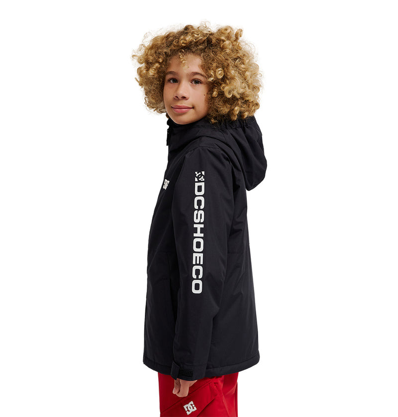A child with curly blond hair is wearing the DC Shoes Basis Youth Jacket—a black, insulated, waterproof jacket featuring white lettering on the sleeve—paired with red pants. The jacket ensures comfort and protection in any weather. The child stands turned slightly to the side and looks at the camera.