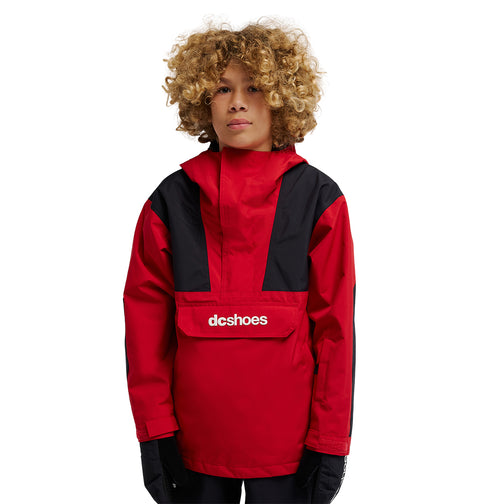 A person with curly blond hair wears the DC Shoes DC-43 Youth Anorak, a red and black insulated snow jacket featuring the “dcshoes” logo on the front pocket, standing against a plain white background.