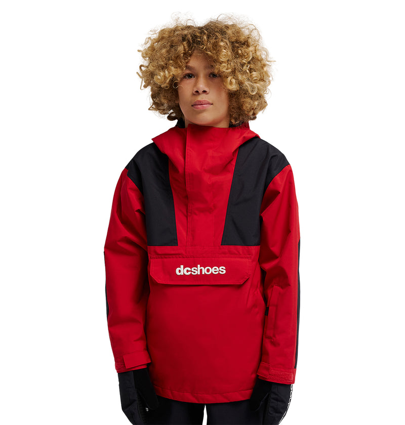 A person with curly blond hair wears the DC Shoes DC-43 Youth Anorak, a red and black insulated snow jacket featuring the “dcshoes” logo on the front pocket, standing against a plain white background.