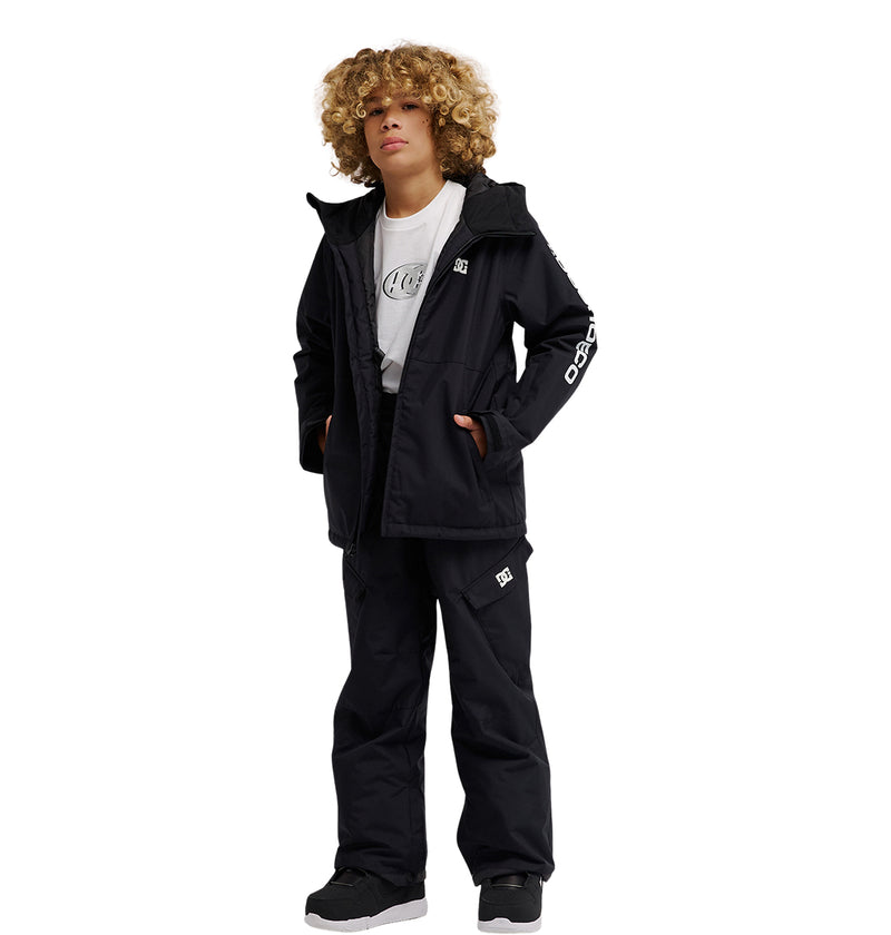 A young person with curly blonde hair stands with hands in pockets against a plain white background, wearing a black jacket, a white t-shirt, black and white sneakers, and DC Shoes Banshee Youth Pants. The outfit combines DC branding with style and insulation for added comfort.