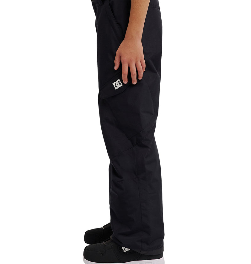A person stands in profile wearing DC Shoes Banshee Youth Pants and black shoes, with one arm resting at their side; the waterproof fabric and added insulation of the pants ensure warmth and comfort.