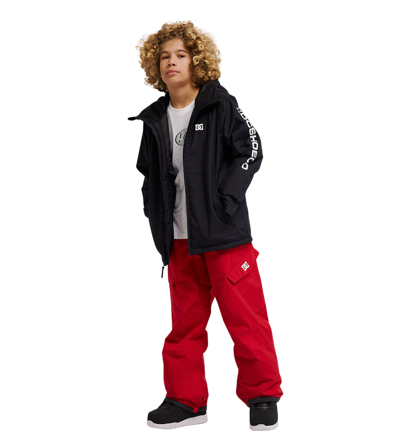 A young person with curly hair wears a black jacket, a white t-shirt, and bright red DC Shoes Banshee Youth Pants with pockets. They stand with their hands in their jacket pockets against a plain white background.