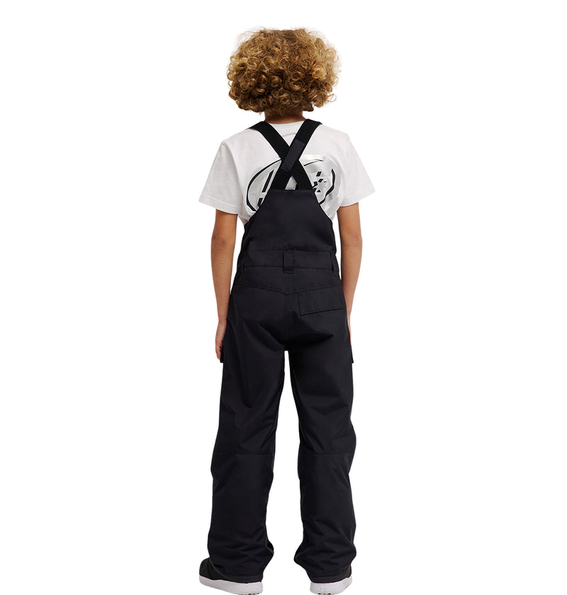 A child with curly hair, wearing the DC Shoes Roadblock Youth Bib over black insulated bib overalls and a white graphic T-shirt, stands facing away from the camera against a plain white background.