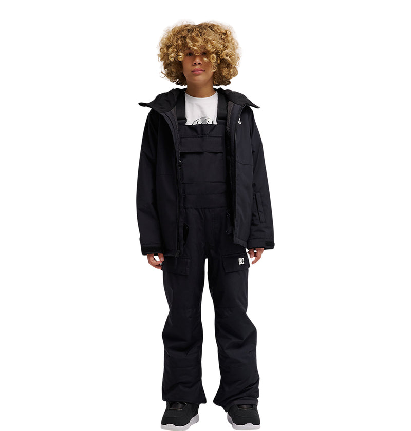 A person with curly blonde hair stands facing forward against a white background, wearing the DC Shoes Roadblock Youth Bib in black over insulated layers, paired with a black jacket, white shirt, and gray sneakers.