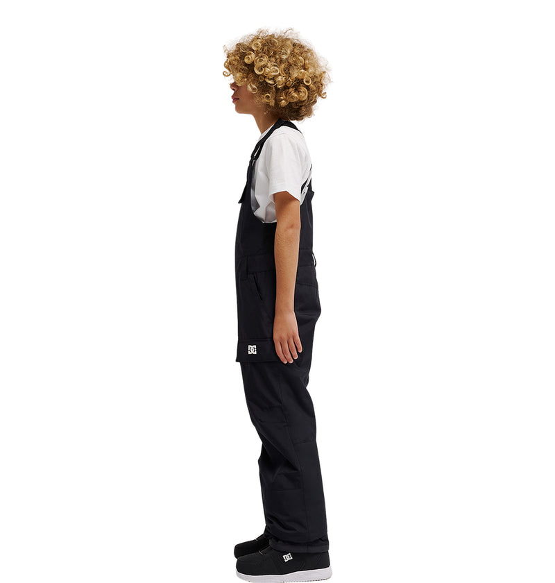 A child with curly blond hair stands in profile against a plain white background, wearing a white T-shirt, black sneakers, and the DC Shoes Roadblock Youth Bib—sleek black insulated overalls.