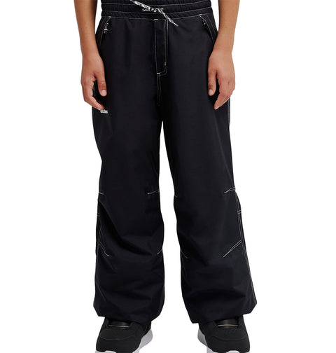A person wearing loose black DC Shoes Trackstar Baggy Youth Pants with white stitching details, featuring a drawstring waist and snow performance waterproofing, paired with black sneakers, stands with hands relaxed at their sides.