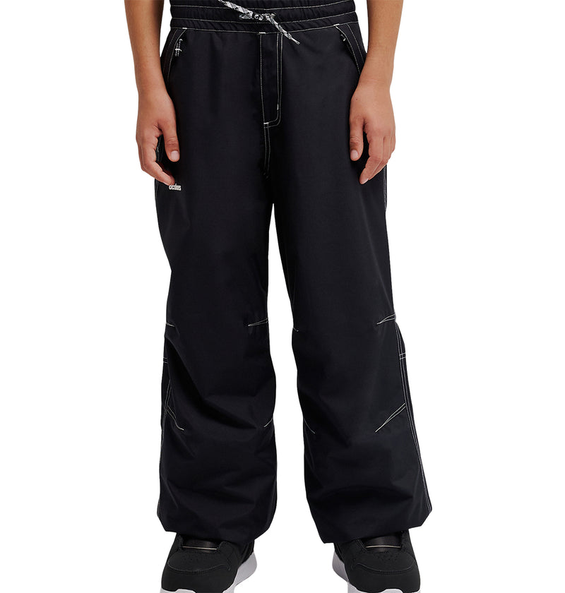 A person wearing loose black DC Shoes Trackstar Baggy Youth Pants with white stitching details, featuring a drawstring waist and snow performance waterproofing, paired with black sneakers, stands with hands relaxed at their sides.