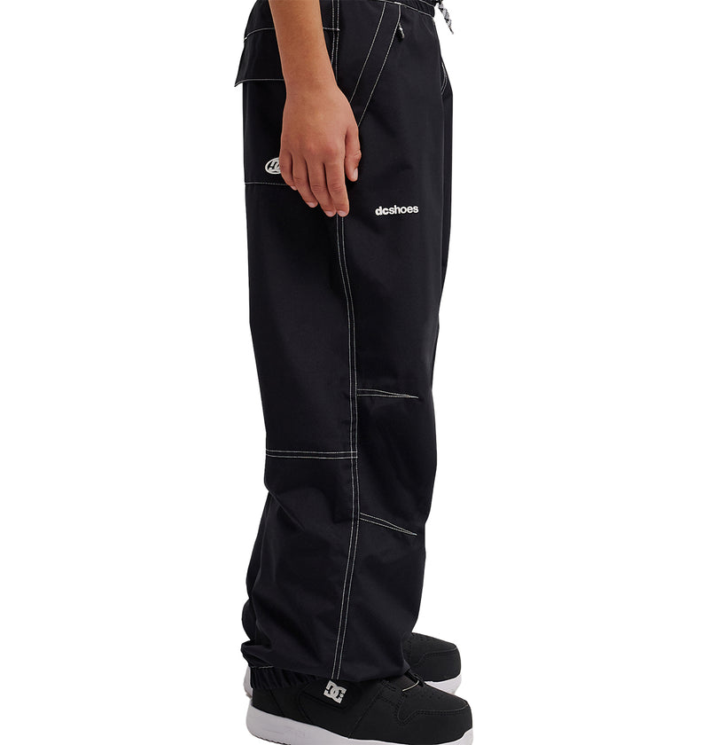 A person is seen standing sideways, showcasing only the lower half of their body, wearing black DC Shoes Trackstar Baggy Youth Pants with white stitching and black skate shoes. Featuring the iconic DC Shoes logo on the thigh, these pants are recognized for their snow performance.