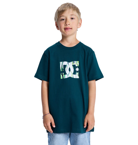A young boy with blond hair wears the Reebok DC Star Filled Ss Boy dark teal graphic t-shirt and light blue jeans, standing and facing the camera against a white background.