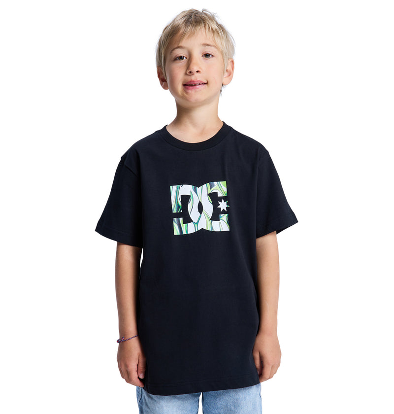 Smiling blond boy wearing the Reebok DC Star Filled Ss Boy loose black T-shirt with a white and green chest logo, standing against a plain white background.