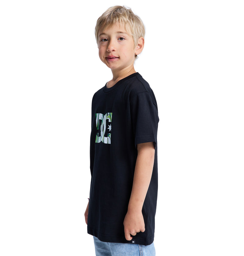 Young boy with light hair wearing the Reebok DC Star Filled Ss Boy black graphic T-shirt and light blue jeans, standing and smiling slightly against a plain white background.