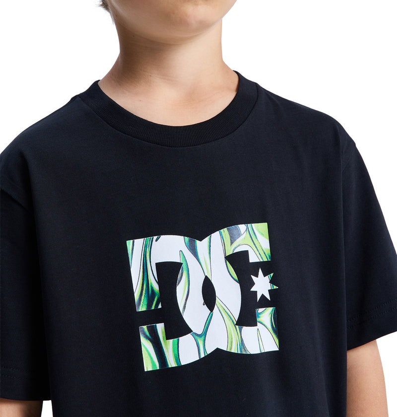 A child wears the Reebok DC Star Filled Ss Boy black T-shirt, featuring a large green and white abstract DC Shoes logo on the front, against a plain white background.