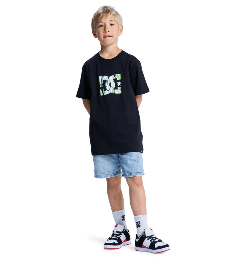 A young boy with blonde hair wears the Reebok DC Star Filled Ss Boy black t-shirt with a green and white graphic, light blue denim shorts, white socks, and black and white sneakers, standing hands behind his back against a white background.