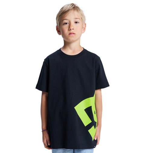 A young boy with short blonde hair faces forward, wearing the Reebok Lanai Youth Hss black t-shirt featuring a large yellow-green side graphic, paired with light blue jeans. The background is plain white.