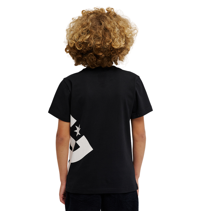 A child with curly blond hair faces away from the camera, wearing the DC Shoes DC Lanai Tee Youth—a relaxed fit black skate tee with a partial white graphic on the lower left—and black pants, standing against a plain white background.