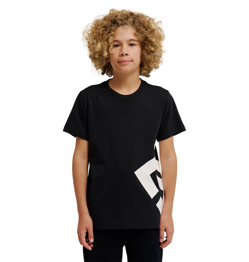 A young person with curly blonde hair wears the DC Shoes DC Lanai Tee Youth, a black relaxed-fit skate shirt with a bold white graphic on one side, standing against a plain white background.