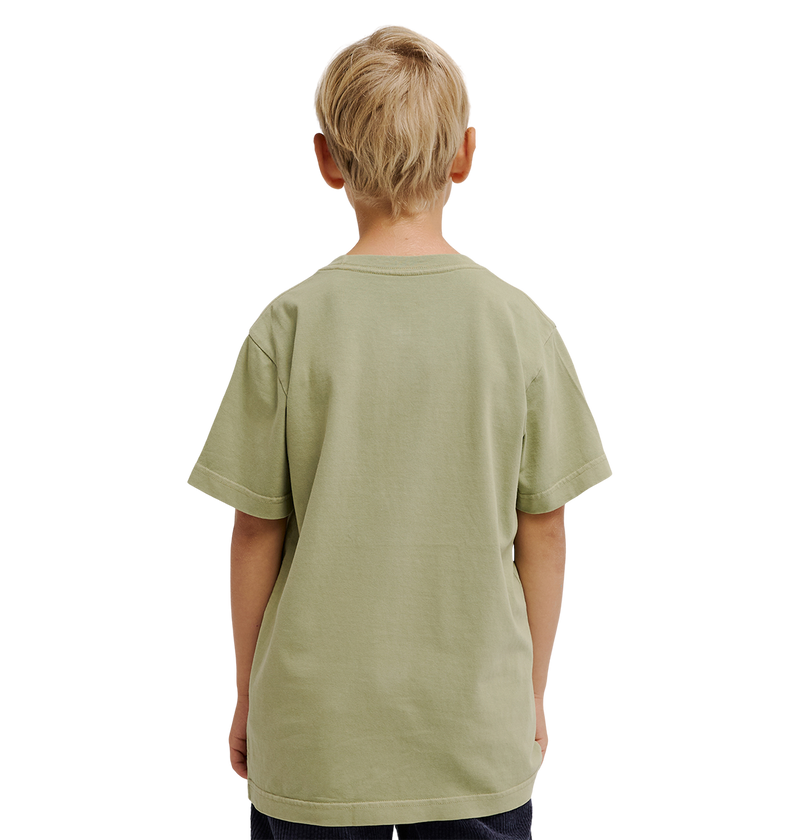 A young boy with short blond hair stands facing away, wearing the DC Shoes DC Speratic Tee Youth in a relaxed fit pale green graphic style and dark pants against a plain white background.