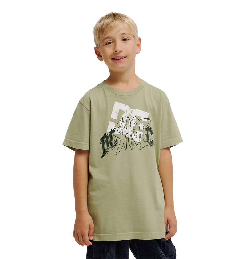 A young boy with short blond hair is wearing the DC Shoes DC Speratic Tee Youth, a relaxed fit light green graphic skate t-shirt featuring heritage-inspired artwork, along with dark pants, as he stands and smiles against a plain white background.