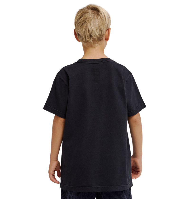 A young boy with short blond hair is standing with his back to the camera, wearing a loose-fitting, plain black DC Speratic Tee Youth by DC Shoes and dark shorts against a light gray background.