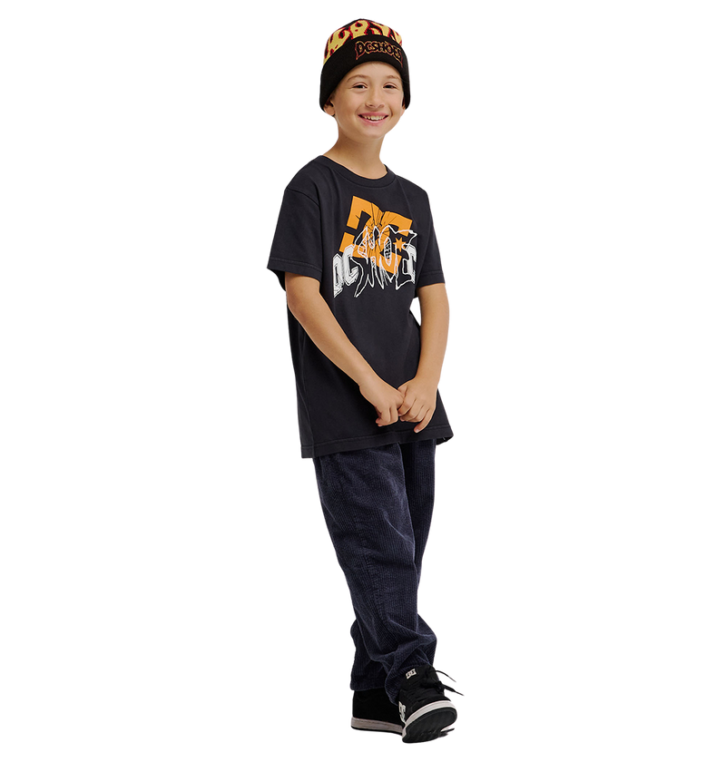 A smiling child stands against a plain white background wearing the DC Shoes DC Speratic Tee Youth in black, paired with dark pants, black sneakers, and a black beanie with orange lettering.