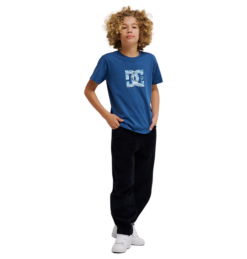A young person with curly blond hair wears the DC Shoes DC Star Filled Tee Youth—a relaxed-fit blue graphic skate t-shirt with bold star artwork—paired with black pants and white sneakers, standing confidently with one hand in their pocket against a plain white background.
