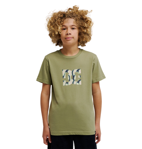 A young person with curly hair is wearing the DC Shoes DC Star Filled Tee Youth, an olive green soft cotton t-shirt with a square camouflage-patterned logo on the chest inspired by iconic DC star artwork, standing against a plain white background.