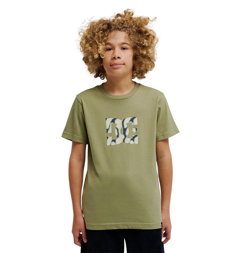 A young person with curly hair is wearing the DC Shoes DC Star Filled Tee Youth, an olive green soft cotton t-shirt with a square camouflage-patterned logo on the chest inspired by iconic DC star artwork, standing against a plain white background.