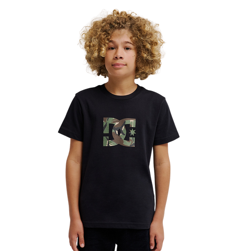 A young person with curly blond hair wears the DC Shoes DC Star Filled Tee Youth, a relaxed fit black graphic skate T-shirt featuring a camo-print DC Shoes star artwork logo on the chest, standing against a plain white background.