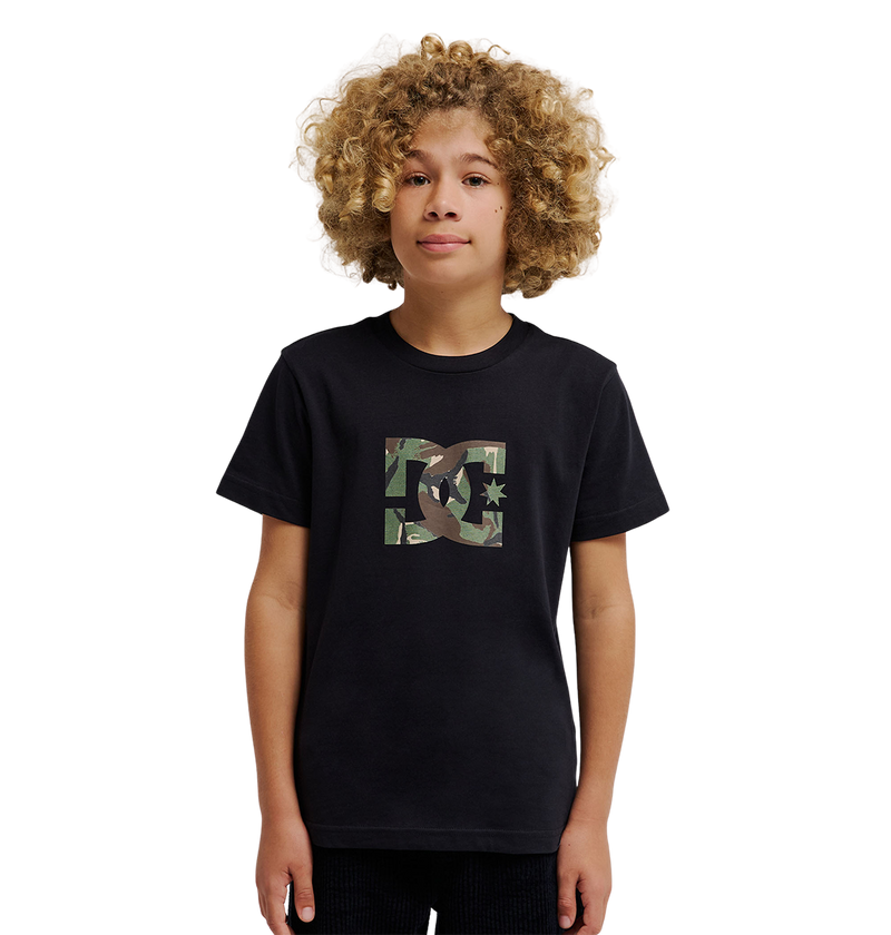 A young person with curly blond hair wears the DC Shoes DC Star Filled Tee Youth, a relaxed fit black graphic skate T-shirt featuring a camo-print DC Shoes star artwork logo on the chest, standing against a plain white background.