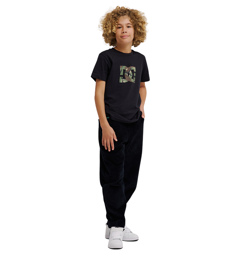 A young person with curly hair stands against a white background, wearing the DC Shoes DC Star Filled Tee Youth—a black graphic skate t-shirt featuring signature DC star artwork—paired with relaxed-fit black pants and white sneakers, one hand in their pocket.