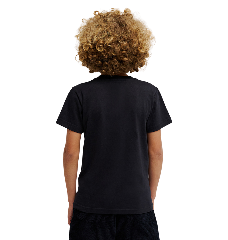 A child with curly blond hair, dressed in a relaxed fit DC Shoes Side Flame Tee Youth featuring subtle flame graphics, and dark shorts, stands with their back to the camera against a light, plain background.