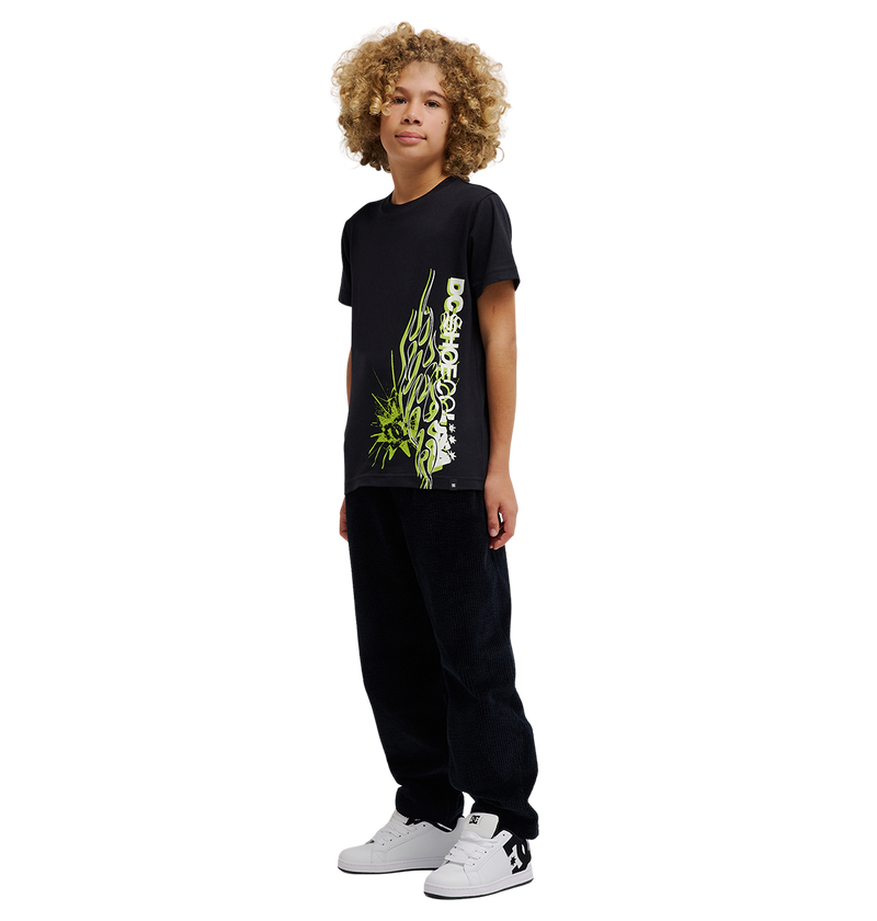 A young person with curly blond hair stands facing forward, wearing a relaxed fit DC Shoes Side Flame Tee Youth in black, featuring green flame graphics, paired with black pants and white sneakers. The background is plain white.