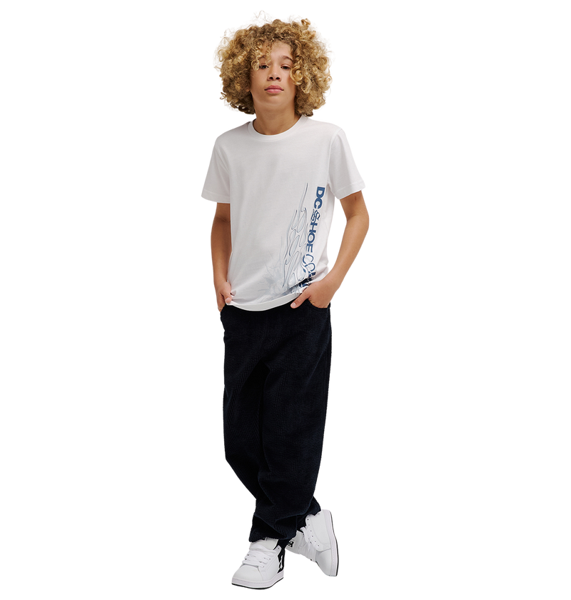 A boy with curly hair wears the DC Shoes DC Side Flame Tee Youth, a white graphic skate tee featuring bold flame artwork, paired with black relaxed fit pants and white sneakers. He stands with his hands in his pockets against a plain white background.