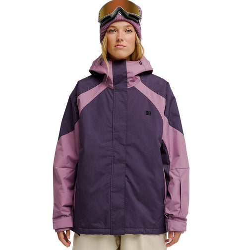 A woman wearing the DC Shoes Everfrost Jacket—a purple and mauve waterproof jacket designed for exceptional snow performance—paired with a pink knit hat and ski goggles, stands facing forward against a white background.