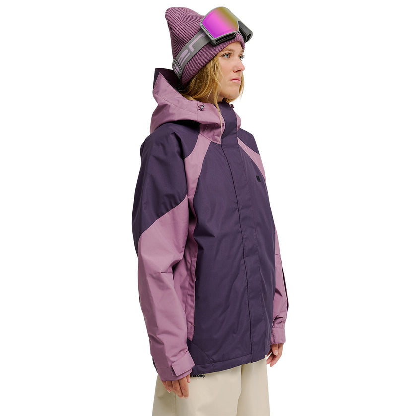 A person wearing a purple and lavender DC Shoes Everfrost Jacket—an advanced waterproof jacket engineered for peak snow performance—paired with beige pants and a knit hat with ski goggles, stands against a plain white background, facing slightly to the right.