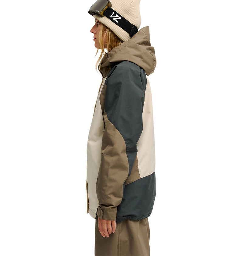 A person wearing the DC Shoes Everfrost Jacket in beige and dark green, paired with brown pants, a white beanie, and ski goggles, stands in profile facing left against a plain white background.