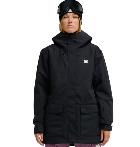 A person wearing a black DC Shoes Cruiser Jacket—an insulated, fully waterproof snow jacket—paired with pink pants, black ski goggles, and a black beanie stands facing forward against a white background.