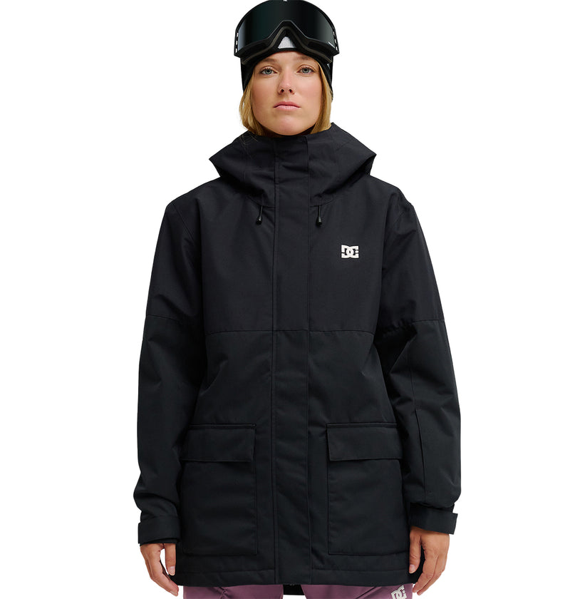A person wearing a black DC Shoes Cruiser Jacket—an insulated, fully waterproof snow jacket—paired with pink pants, black ski goggles, and a black beanie stands facing forward against a white background.