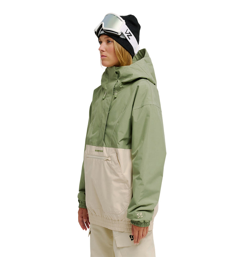 A woman wears the DC Shoes Chalet Anorak—a green and beige insulated ski jacket—paired with beige pants, a black beanie, and ski goggles resting on top. She stands against a white background, looking to the side. This look is perfect for enhanced snow performance and reliable waterproofing.