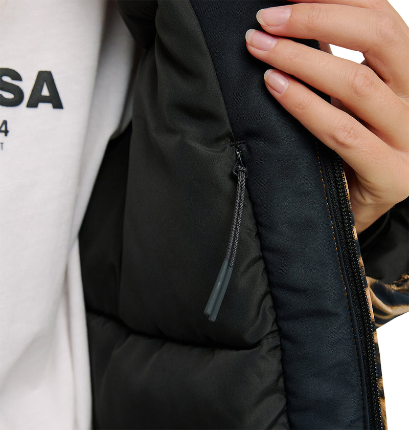 A close-up of a person’s hand holding open the black DC Shoes Aura Puffer Jacket, revealing an inside zippered pocket. Part of a white shirt with black text is also visible, emphasizing the jacket’s breathable protection.