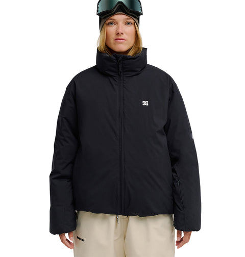 A person stands facing forward against a white background, wearing the DC Shoes Aura Puffer Jacket in black, beige pants, and ski goggles on their head.