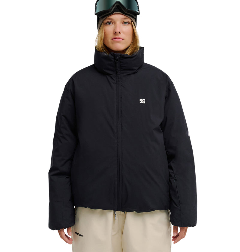 A person stands facing forward against a white background, wearing the DC Shoes Aura Puffer Jacket in black, beige pants, and ski goggles on their head.