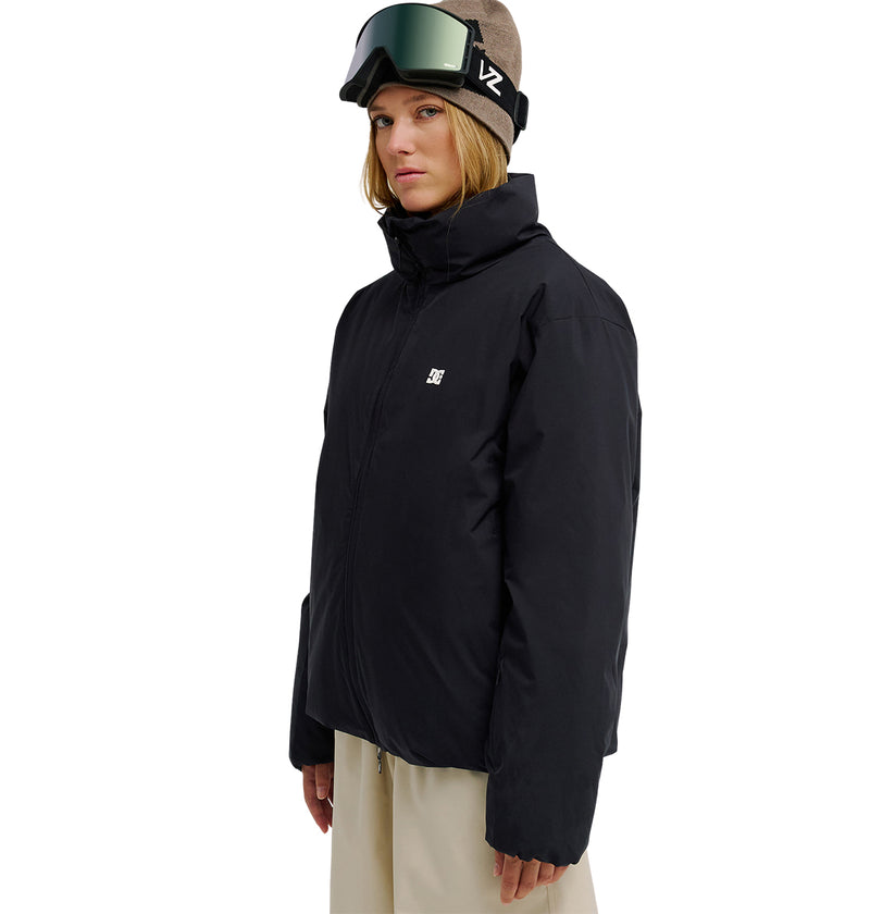 A person wearing the DC Shoes Aura Puffer Jacket in black, paired with beige pants and a brown beanie topped with dark ski goggles, stands against a plain white background while looking slightly to the side.