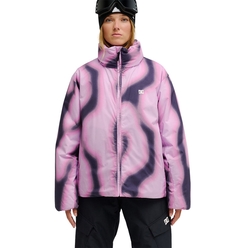 A person is wearing the DC Shoes Aura Puffer Jacket, featuring a pink and black wavy pattern. The waterproof jacket offers breathable protection for enhanced snow performance. The individual, dressed in black pants with ski goggles on their head, is standing against a white background.