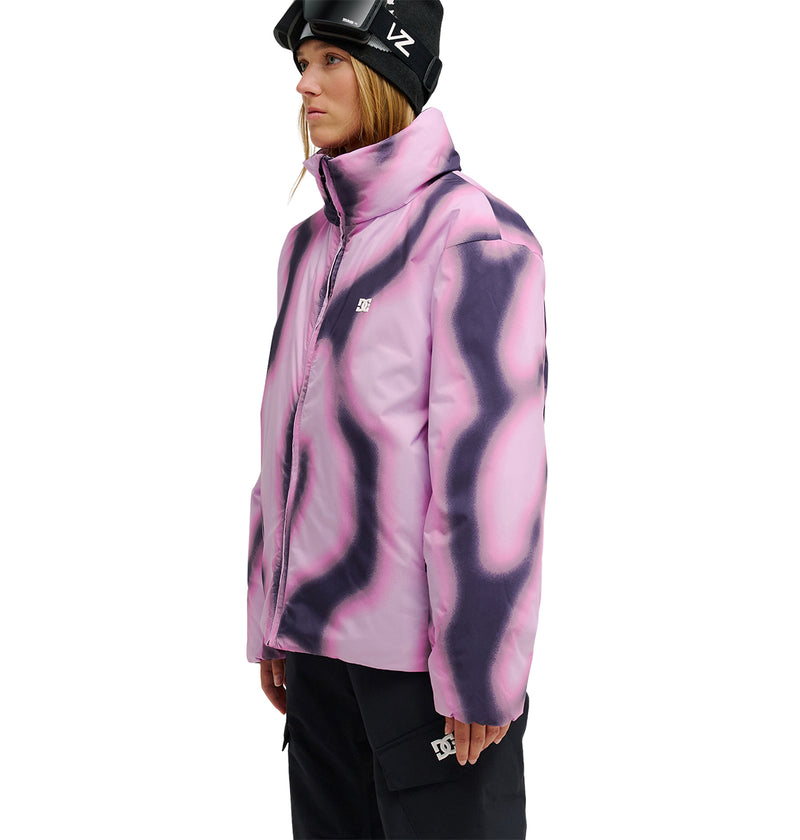 A person wearing the DC Shoes Aura Puffer Jacket, featuring a pink and purple wavy pattern, along with black pants and ski goggles, stands against a plain white background facing left.